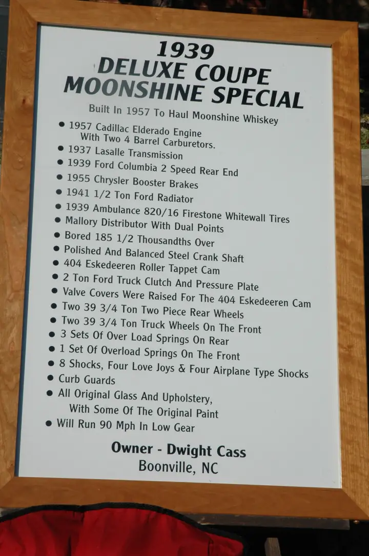A framed white placard headed "1939 Deluxe Coupe Moonshine Special" lists the car's parts and modifications in a bulleted list and names the owner and his hometown of Boonville, NC.