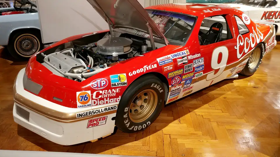 A red-and-white No. 9 NASCAR Ford Thunderbird covered in sponsor decals, with its hood raised to show the engine, sits on a polished parquet museum floor beside other cars.