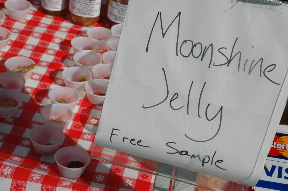 A hand-lettered sign reading "Moonshine Jelly, Free Sample" stands on a red-and-white checked tablecloth next to small paper sample cups and jars; a card-payment sticker shows at the right edge.