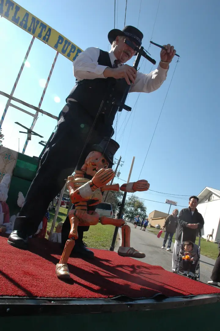 Low-angle view of a man in a black hat, vest and tie working a large wooden skeleton-like marionette on a red-carpeted stage under a yellow banner reading "Atlanta Pupp..."; spectators, including a child in a stroller, watch from the street behind.