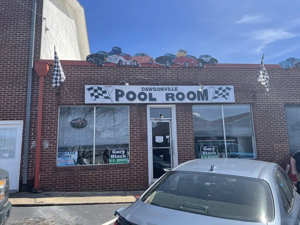 A brick storefront with a "Dawsonville Pool Room" sign flanked by checkered flags, a row of model race cars along the roofline, glass windows with posters and campaign signs, and a silver car parked in front.